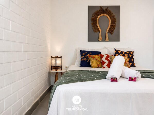 A hotel room with a white brick wall, a bed with a green polka dot bedspread, multiple colorful pillows, a wall-mounted artwork, and a small table with a lamp.