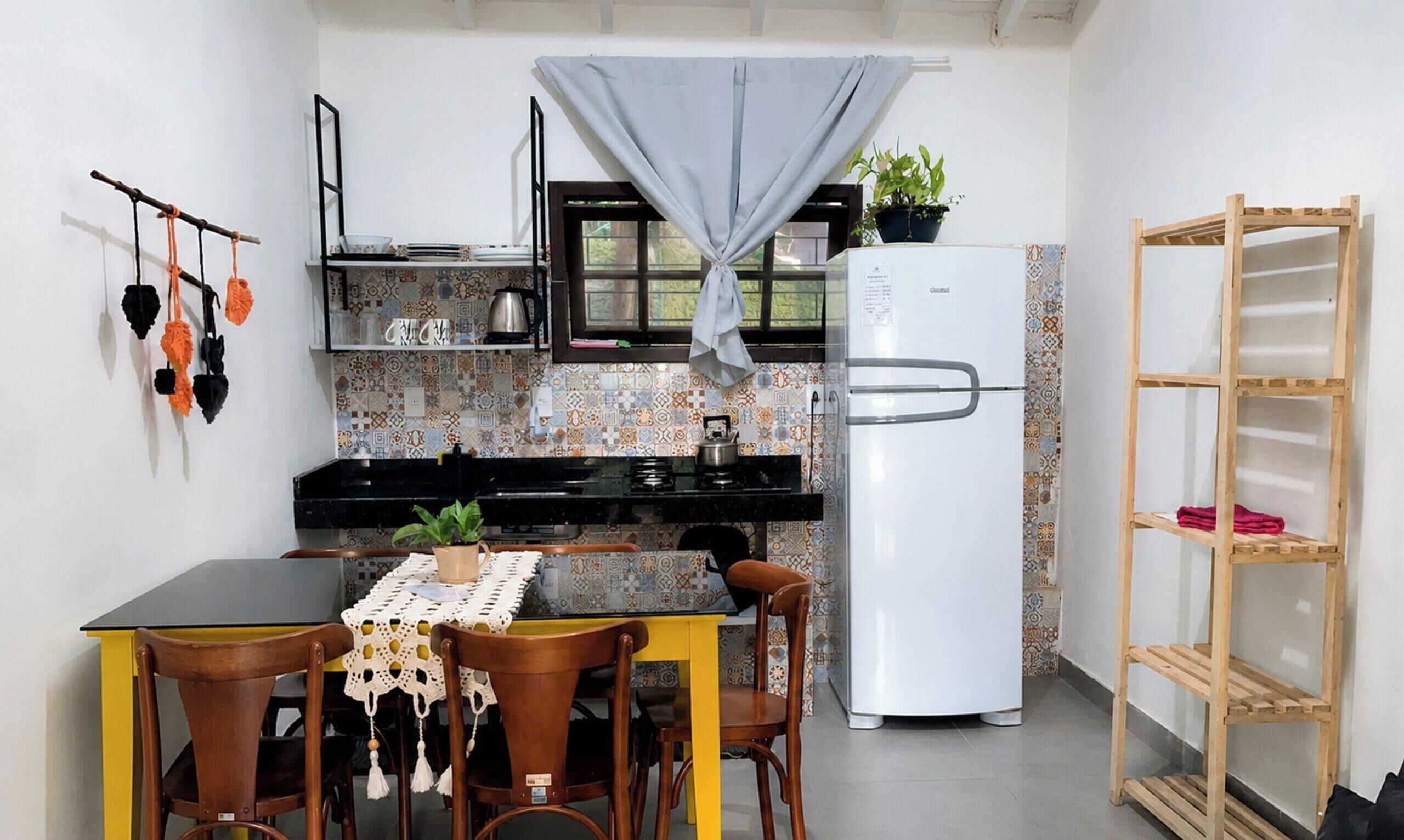 A compact kitchen with a refrigerator, a dining table with chairs, and a small shelf unit.