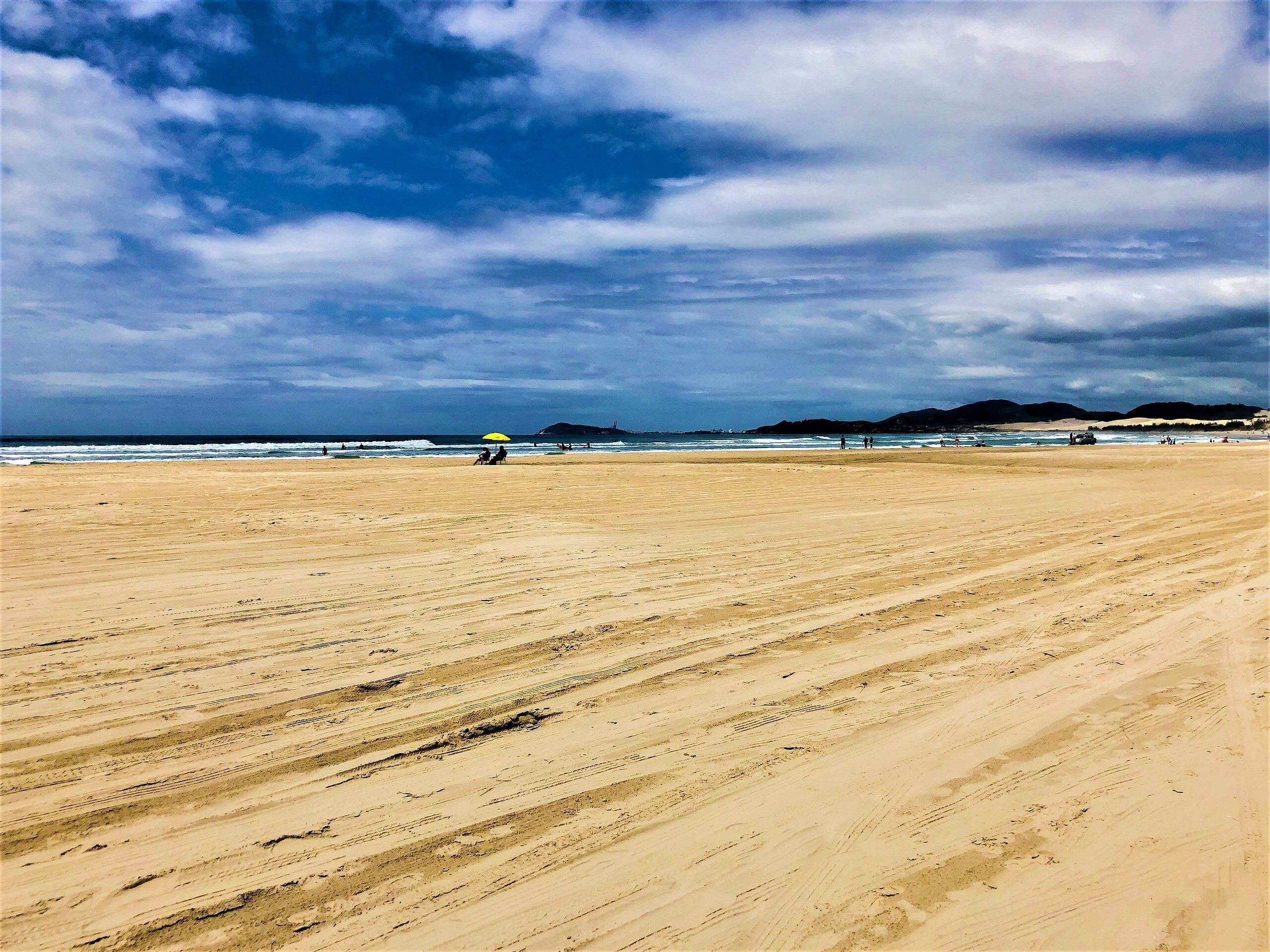 Uma extensa praia de areia com algumas pessoas e um guarda-sol amarelo ao longe.