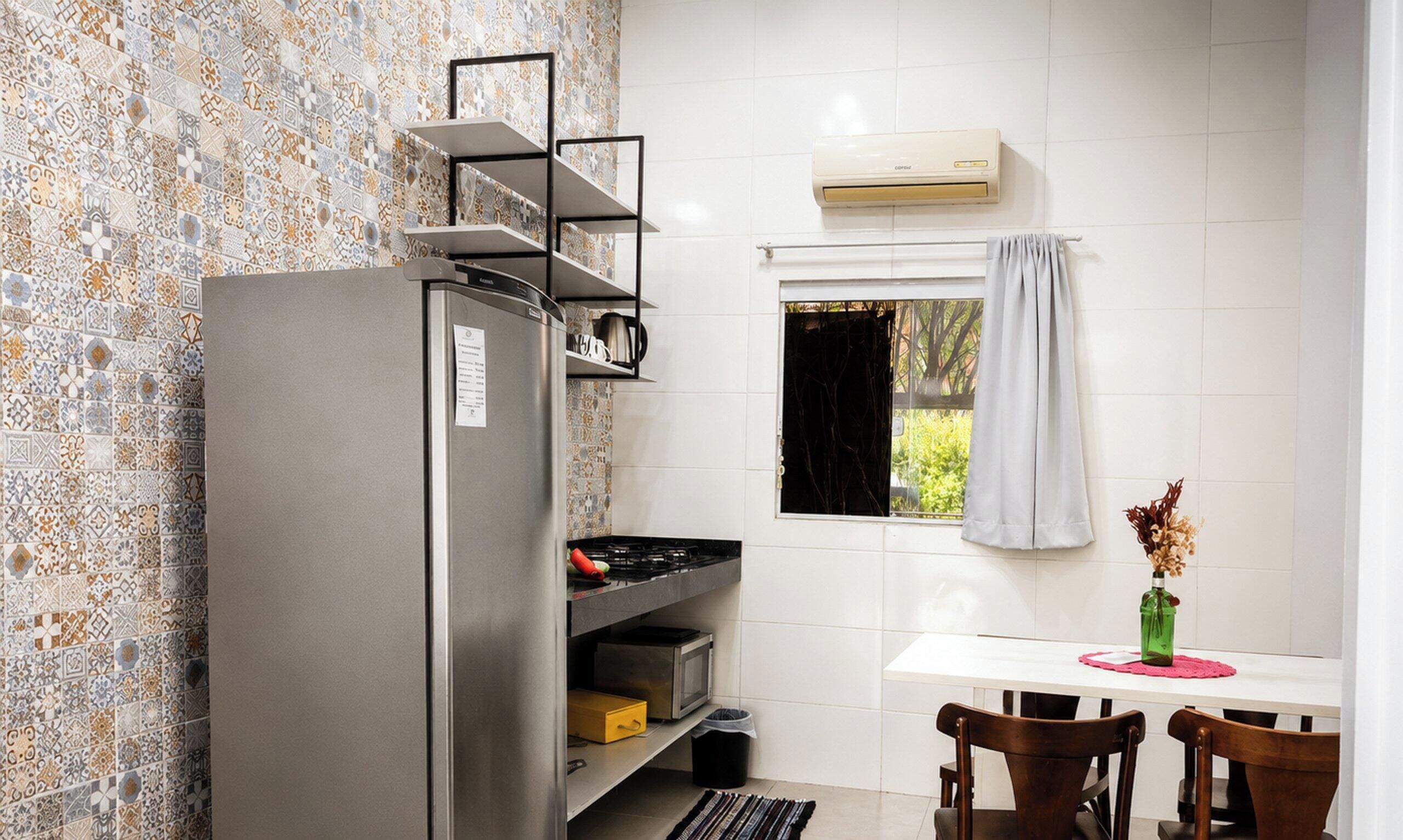 A compact kitchen with a refrigerator, a window with curtains, a small dining table with chairs, and a decorative vase.