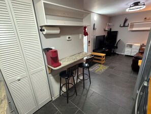 Cozinha compacta com bancada, cafeteira, televisão e área de estar com banquetas.
