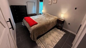 Ett sovrum med en säng, ett nattduksbord, en byrå, en garderob, en taklampa och ett fönster med gardiner.
