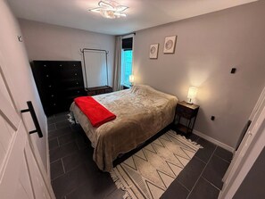 Quarto com cama, mesa de cabeceira, cômoda, guarda-roupa, luminária no teto e janela com cortinas.