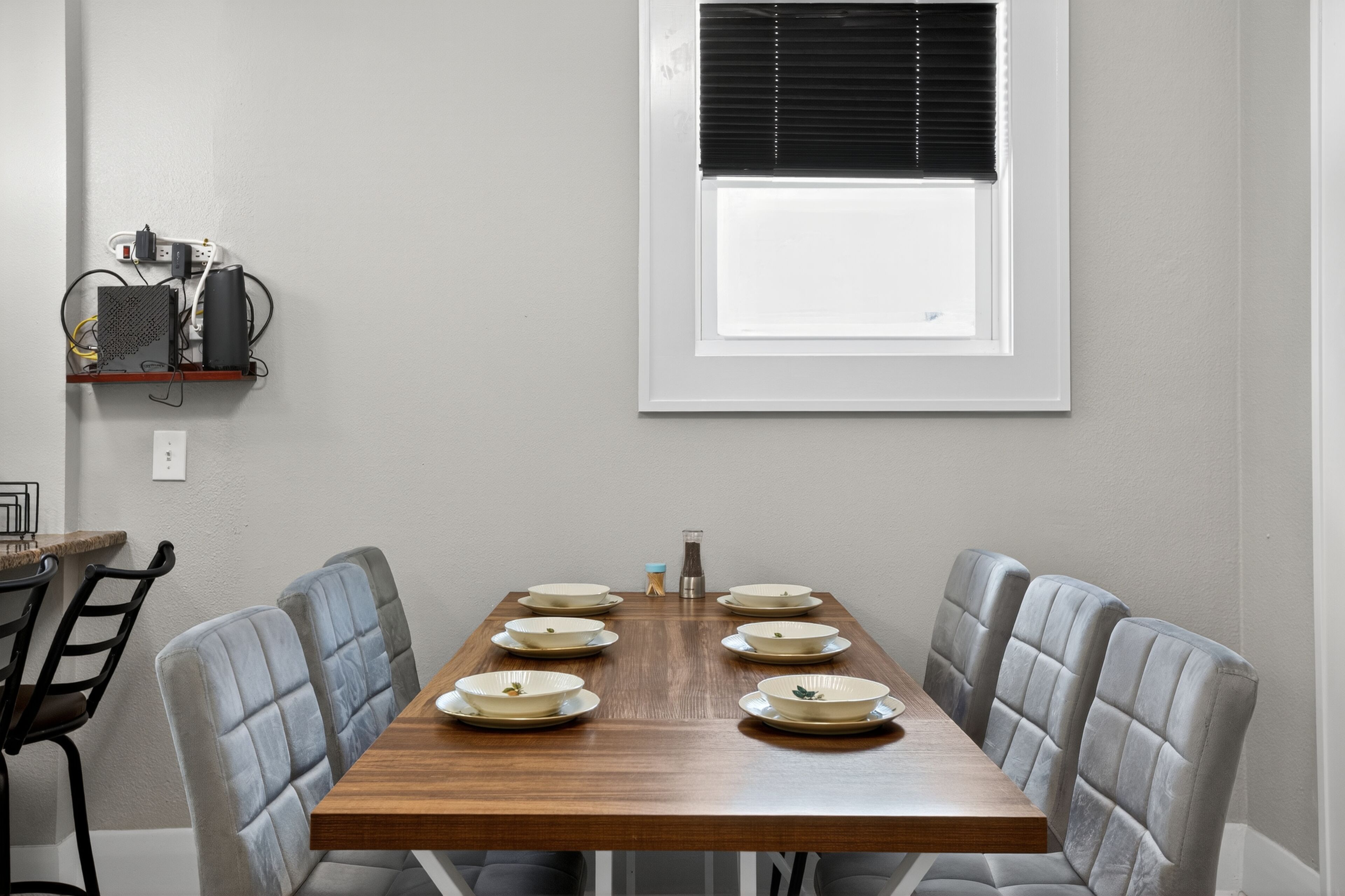 Una mesa de comedor con seis platos y una jarra, rodeada por seis sillas, en una habitación con ventana e interruptores en la pared.