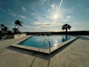 A rectangular swimming pool with a metal ladder, surrounded by a tiled deck, palm trees, and a clear sky. - Beachfront 1BR condo with pool, WiFi, and Gulf views. (Marco Island)