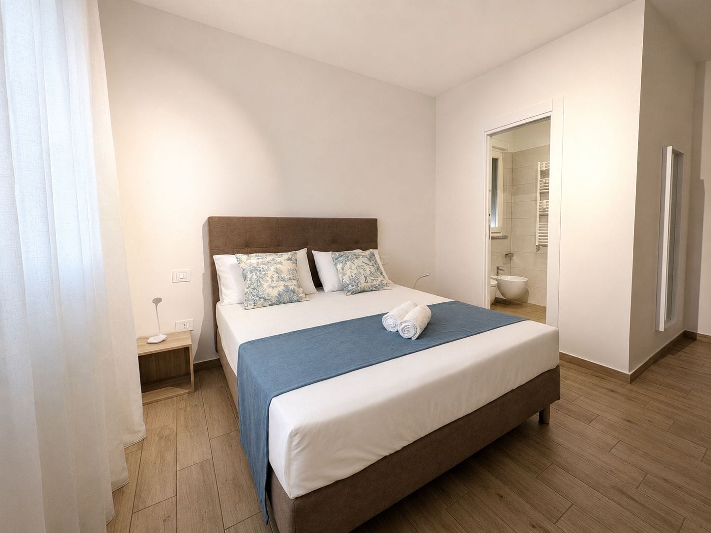 Un dormitorio con cama, mesita de noche, lámpara de noche y un baño visible a través de una puerta abierta.