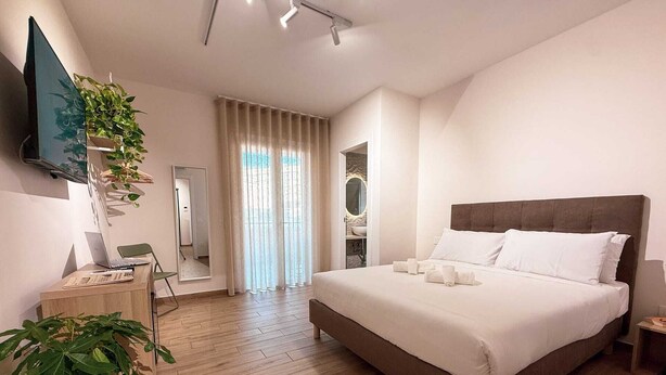 Una habitación de hotel moderna con una cama grande, un escritorio con una computadora portátil, un televisor montado en la pared y una planta en maceta.