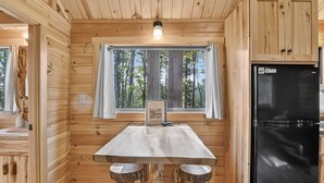 Ein Holzblockhaus-Innenraum mit Esstisch, zwei Barhocker, einem Kühlschrank und einem Fenster mit Vorhängen.