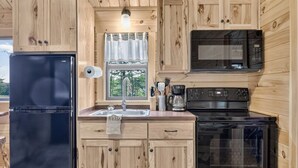 Eine kleine Küche mit Holzschränken, einem schwarzen Kühlschrank, einer Mikrowelle, einem Herd und einem Fenster mit Vorhängen.