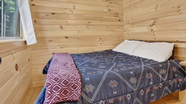 Ein Zimmer in einer Holzhütte mit einem Bett, einem Fenster mit Vorhängen und Blick auf Bäume.