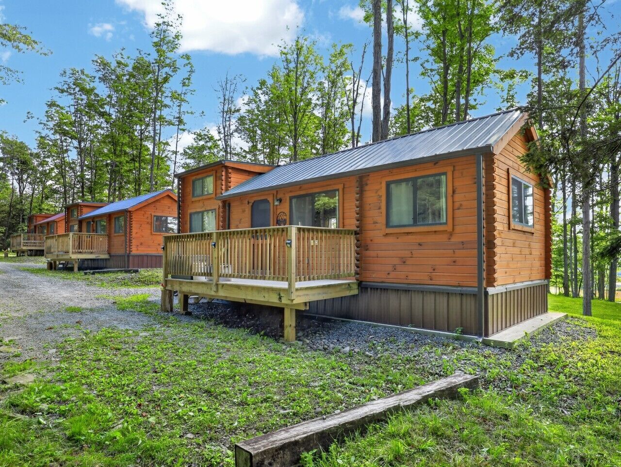 A row of wooden cabins with a deck, surrounded by trees and a gravel path.