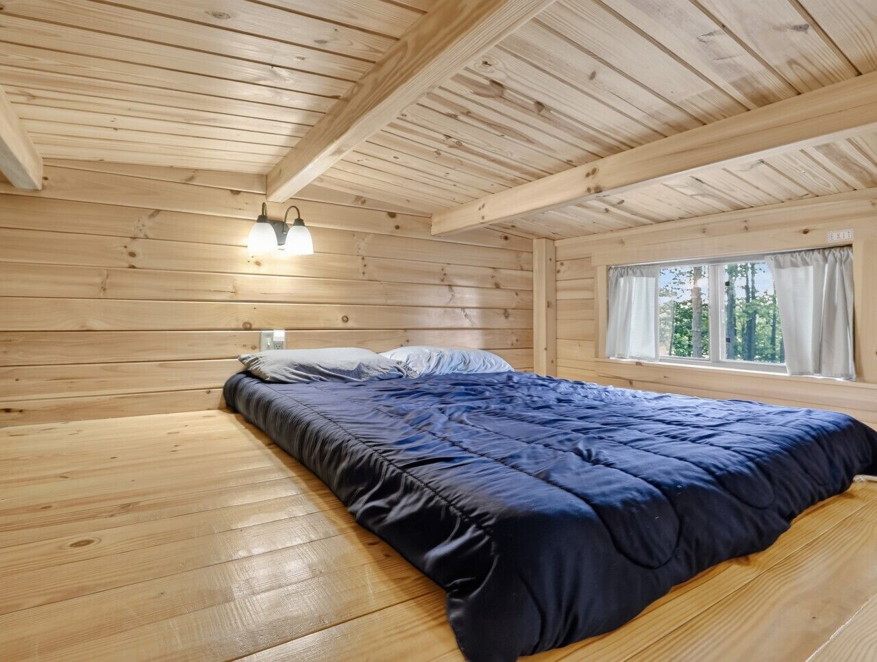 A wooden cabin bedroom with a bed, a window with curtains, and a light fixture.