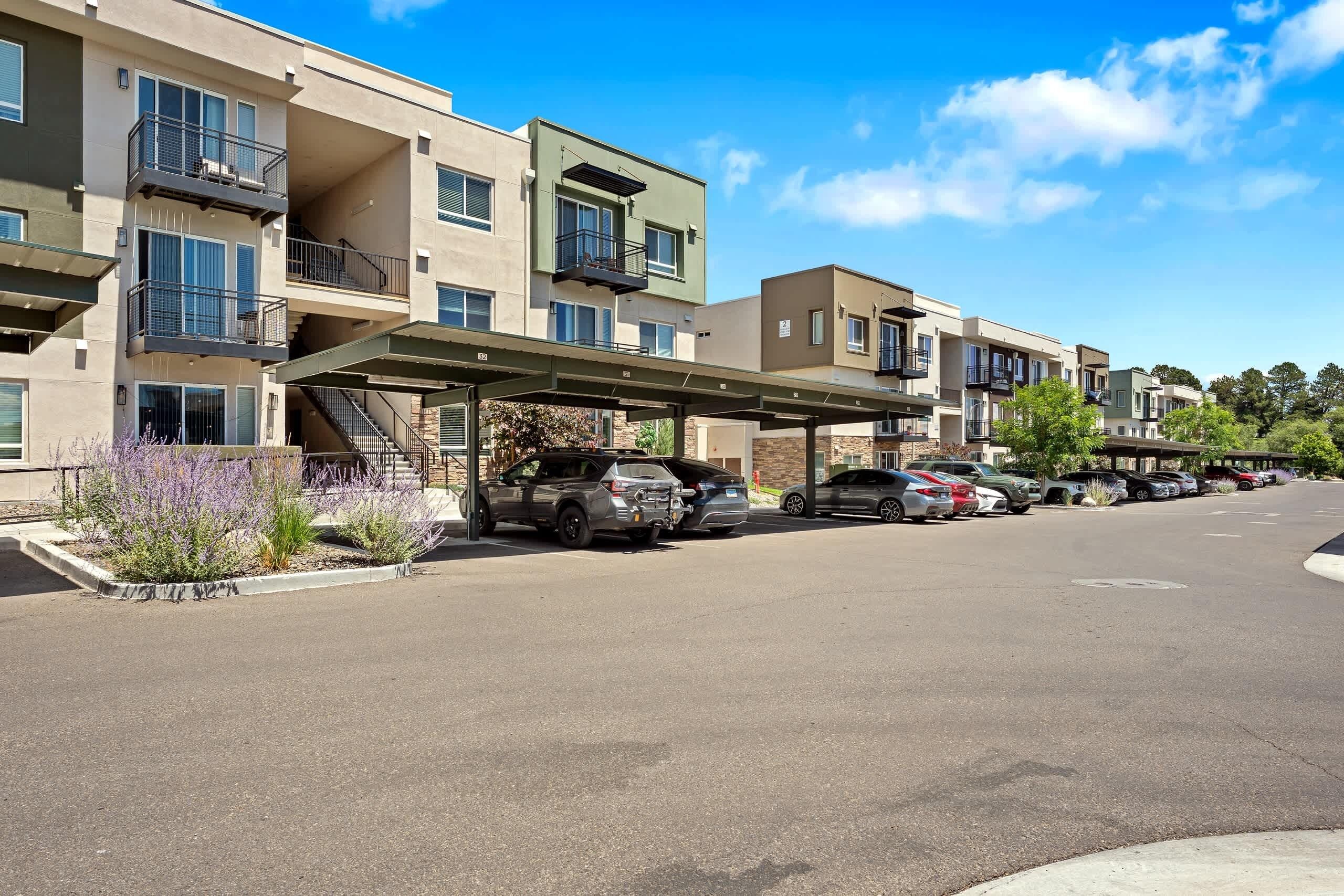 A multi-story residential building with balconies, a covered parking area, and a row of parked cars.