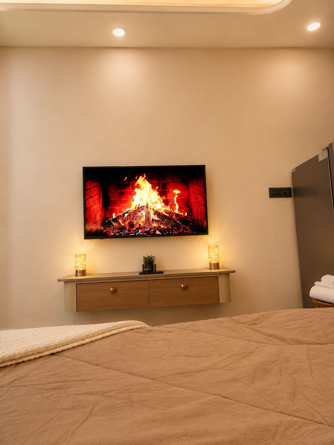 Ein Schlafzimmer mit einem Bett, einem an der Wand montierten Fernseher, einem Nachttisch mit zwei Lampen und einem Kühlschrank.