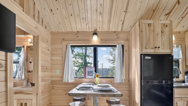 Interior de una cabaña de madera con una mesa de comedor, cuatro taburetes, un refrigerador negro y una ventana con cortinas.