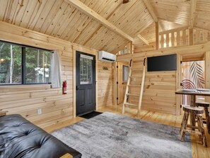 Ein Blockhaus-Interieur mit einer schwarzen Tür, einem Feuerlöscher, einem Fenster, einer Holzbalkendecke und einem grauen Teppich.