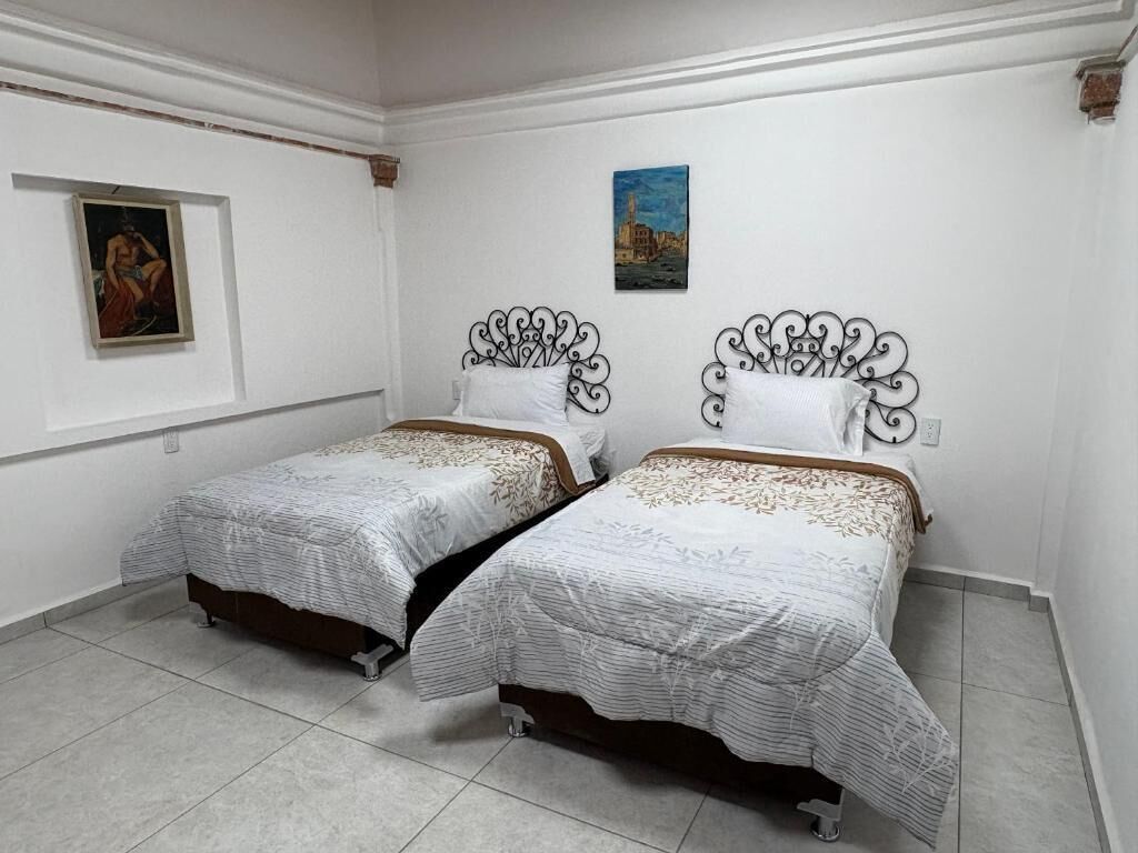 A room with two beds, white walls, a painting on the left, and a small picture on the wall above the beds.