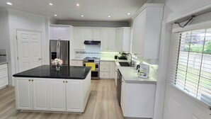A modern kitchen with white cabinetry, a central island, a stainless steel refrigerator, and a window with blinds. - Bright & Spacious Estate (Surrey)
