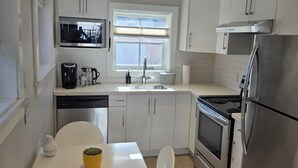 A modern kitchen with white cabinets, stainless steel appliances, a window with blinds, and a dining area with a white table and chairs. - Private Comfortable Guest House (Vancouver)