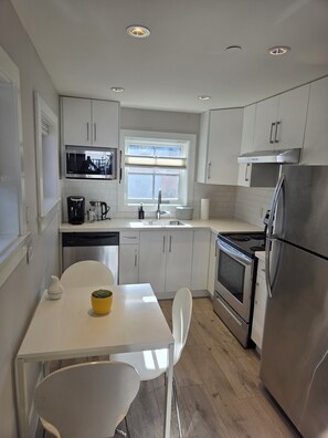 A modern kitchen with white cabinets, stainless steel appliances, a window with blinds, and a dining area with a white table and chairs. - Private Comfortable Guest House (Vancouver)
