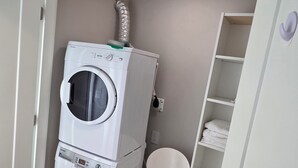 A compact laundry room with stacked washing machines, a chair, and a shelving unit. - Private Comfortable Guest House (Vancouver)