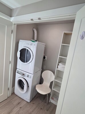 A compact laundry room with stacked washing machines, a chair, and a shelving unit. - Private Comfortable Guest House (Vancouver)