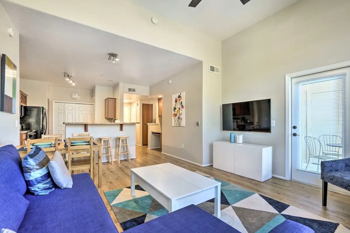 A modern living room with a sofa, coffee table, and a TV. There is a kitchen area with cabinets and a dining table.