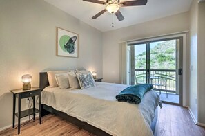 Ein Schlafzimmer mit Bett, Nachttischlampen, Deckenventilator, einem Fenster mit Blick auf Grünflächen und einem Balkon mit Gartenmöbeln.