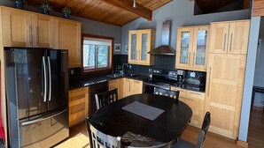 A modern kitchen with wooden cabinets, a stainless steel refrigerator, a dining table with chairs, and a window with a view of trees. - Invermere Getaway with Stunning Views and Hot Tub! (Invermere)