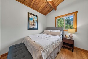 A bedroom with a bed, a bench, a nightstand, a lamp, a framed picture, and a window with a view of trees.