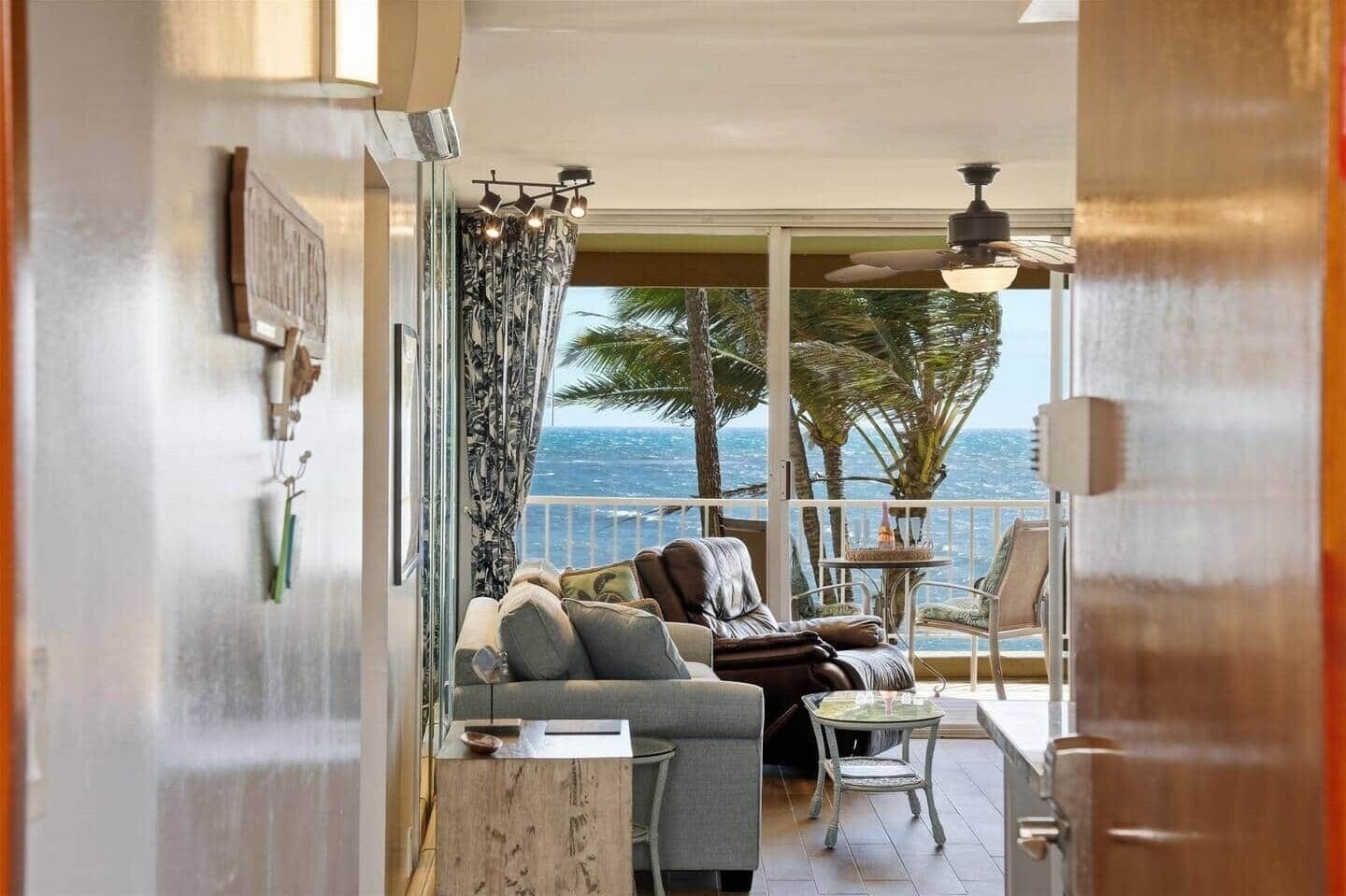 A balcony with a sofa, chairs, and a table, overlooking a view of palm trees and the ocean.