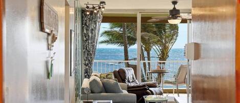 A balcony with a sofa, chairs, and a table, overlooking a view of palm trees and the ocean.