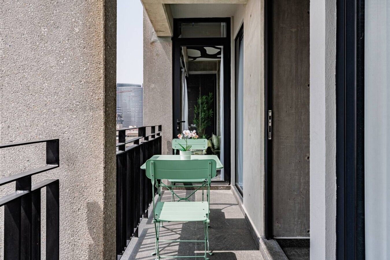 Un balcone stretto con un tavolo e sedie in metallo verde, una pianta in vaso e vista su un edificio sullo sfondo.