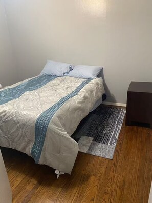 Ein Einzelbett mit einem gemusterten Überwurf, ein kleiner Teppich und ein Nachttisch mit einer Schachtel darauf.