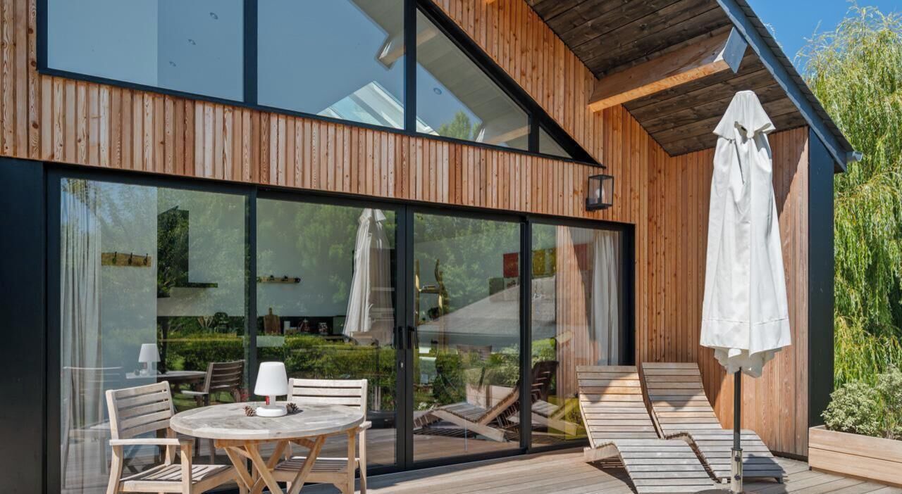 A modern wooden building with large glass windows, outdoor seating, and a patio area.
