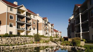 Un immeuble résidentiel à plusieurs étages, doté de balcons, d’un mur de soutènement en pierre et d’un petit étang.