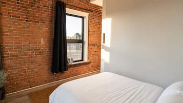Une chambre avec un mur en briques, une fenêtre avec un rideau, un lit avec une literie blanche et un plancher en bois.