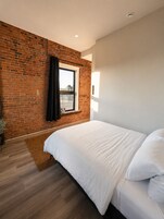 Un dormitorio con pared de ladrillo, ventana con cortina, cama con ropa blanca y piso de madera.