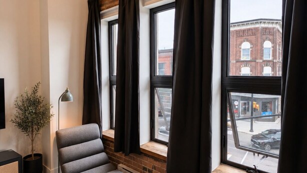 Un salón moderno con un sofá, una planta en maceta, una lámpara de pie y ventanas grandes con cortinas.