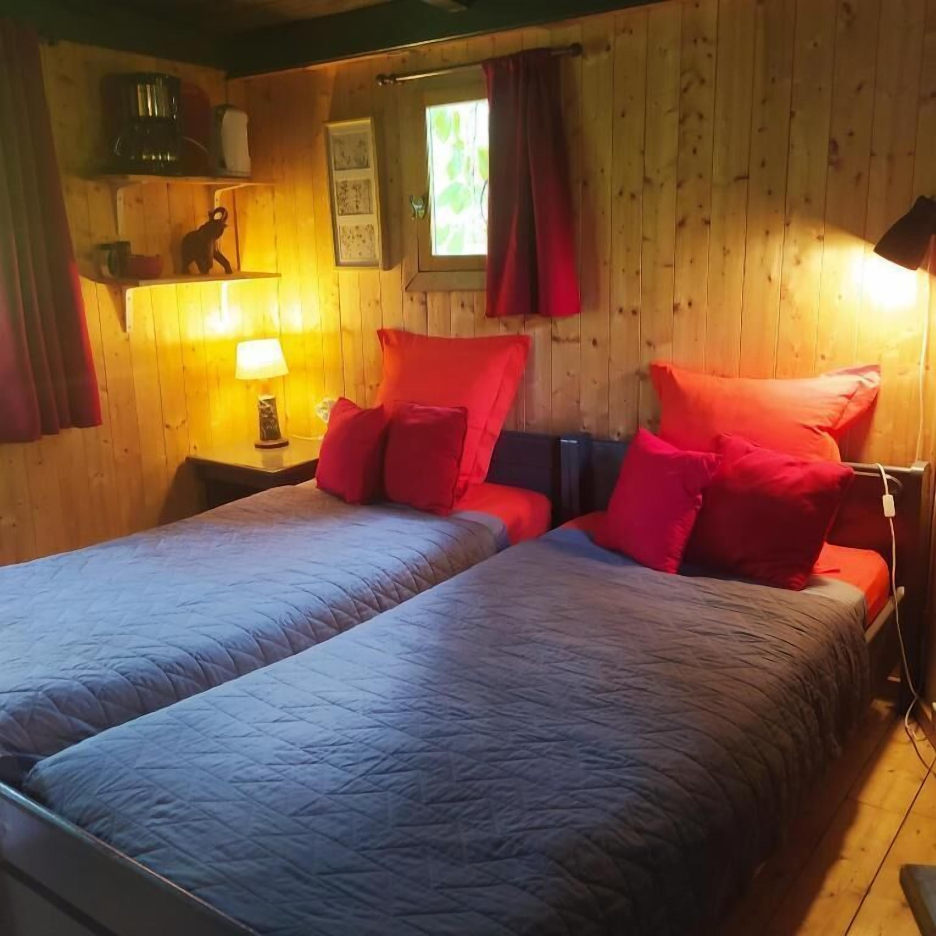 Une chambre avec deux lits, des murs en bois, une fenêtre avec des rideaux, une lampe et une petite étagère avec quelques objets.