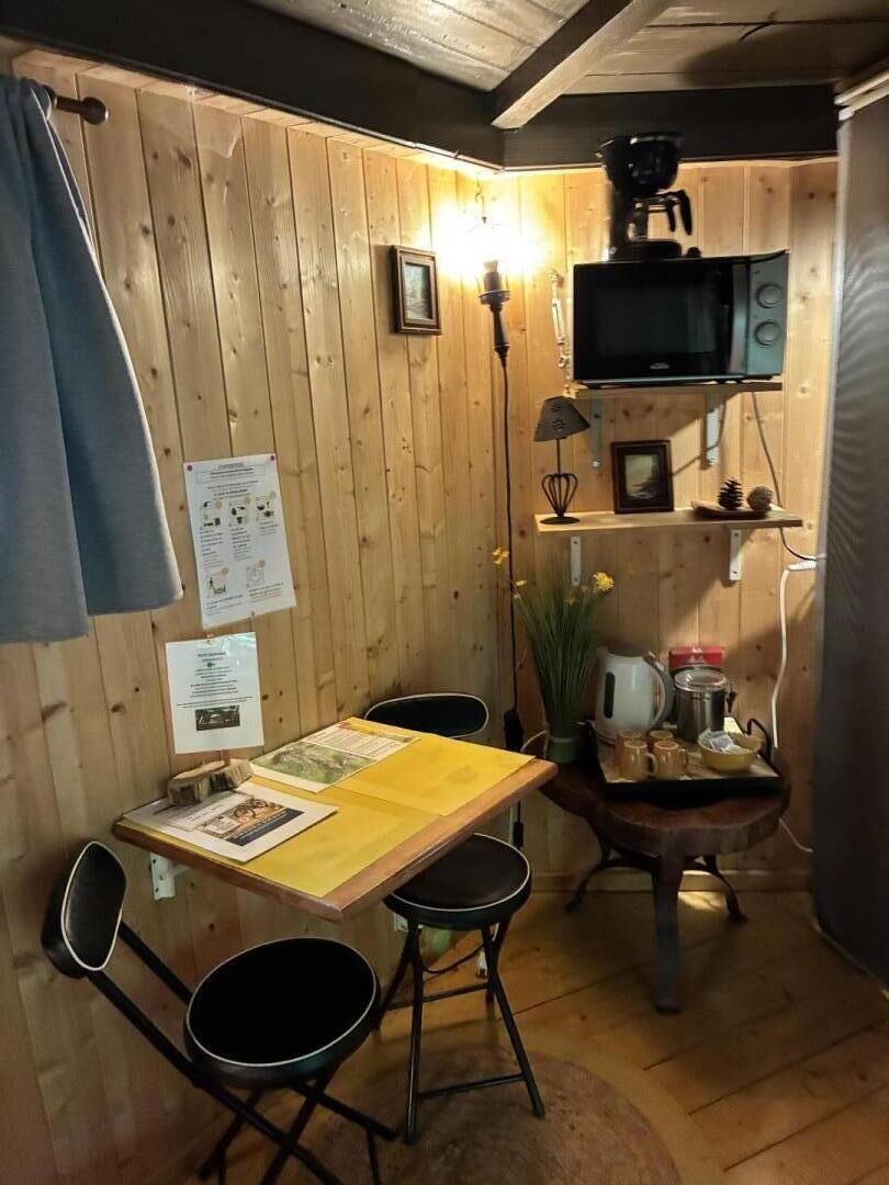 Een kleine houten kamer met een tafel, stoelen, een magnetron en een plank met verschillende spullen.