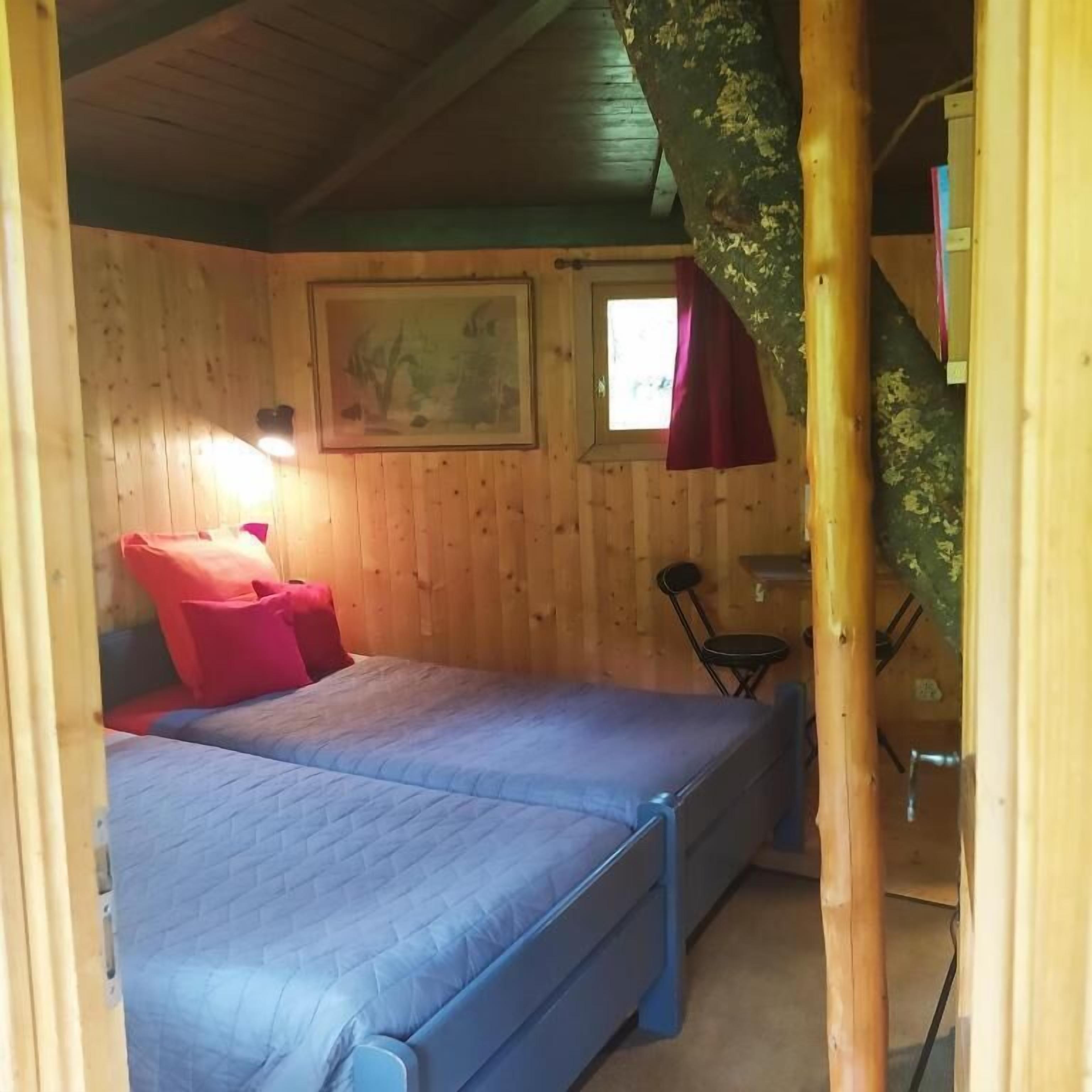 Ein Zimmer mit Holzwänden, einem Bett mit blauer Bettwäsche, einem kleinen Tisch und einem Stuhl.