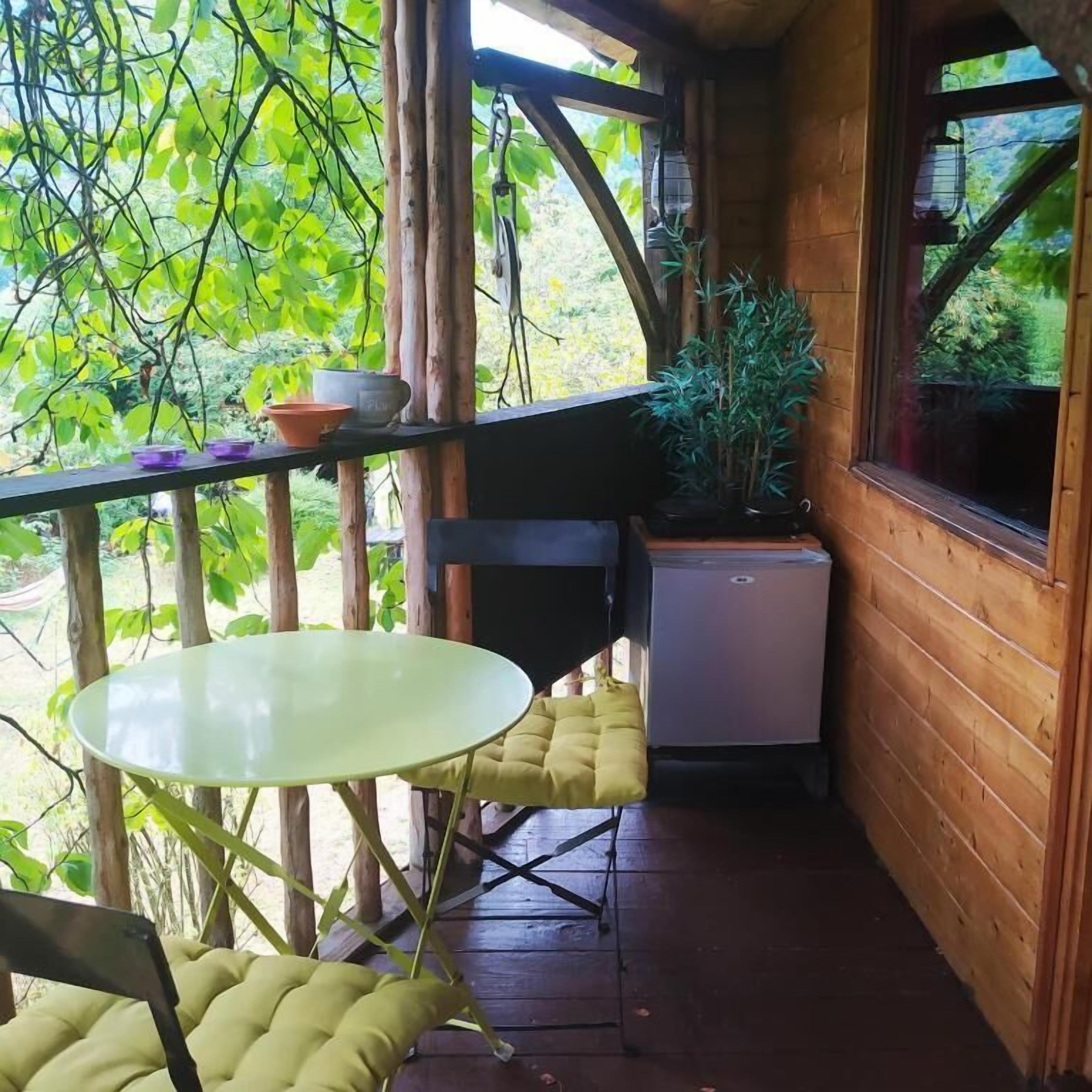 A wooden cabin with a balcony, a table, chairs, a potted plant, and a small refrigerator.