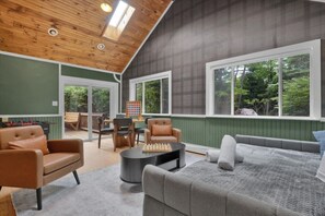 Une pièce spacieuse avec un plafond en bois, un mur vert et une grande fenêtre donnant sur les arbres. On y trouve un canapé, une table basse et une table d’échecs avec un jeu déjà installé.