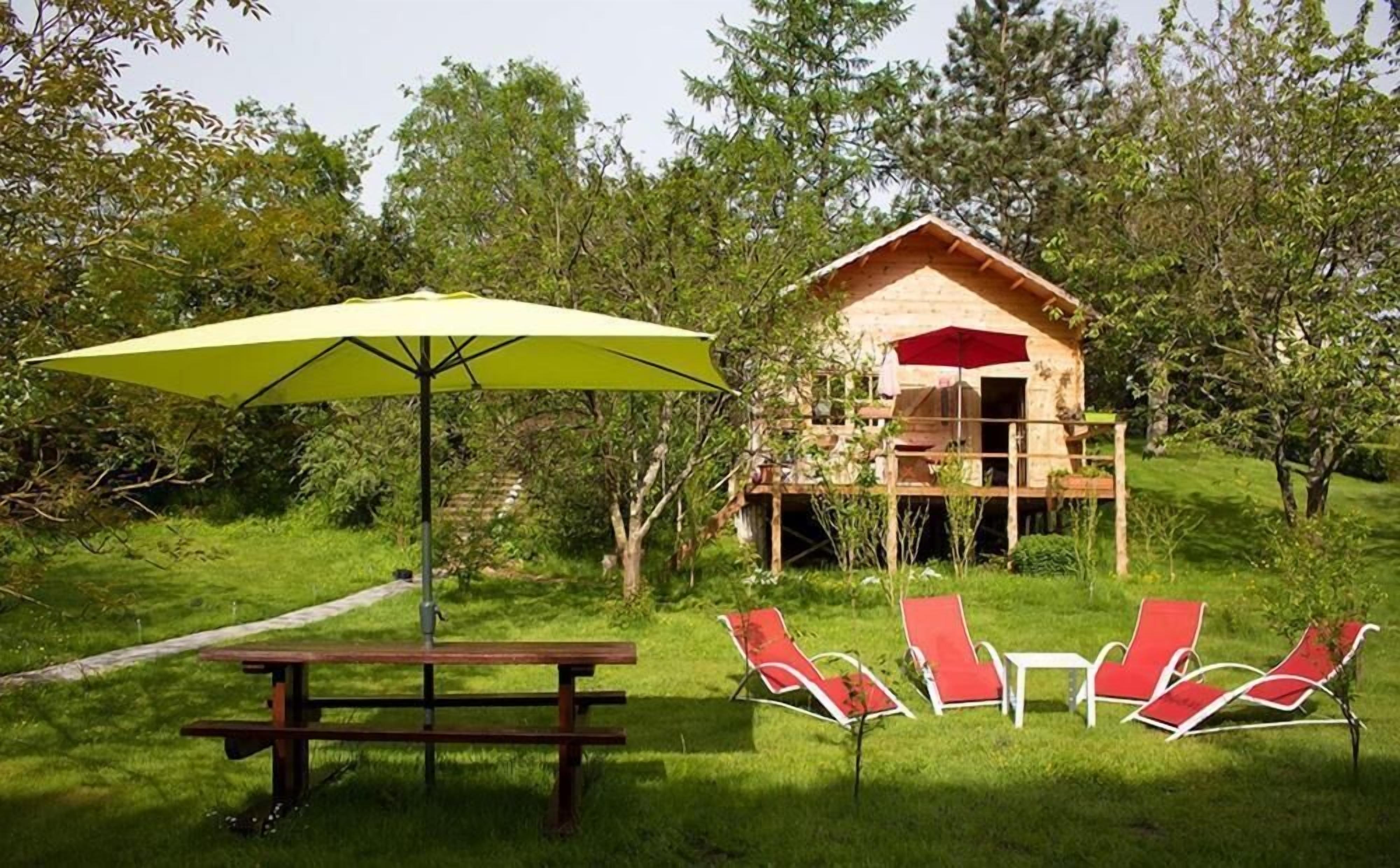 Une cabane en bois dotée d’une terrasse, entourée de verdure, avec une table de pique-nique et des chaises longues rouges.