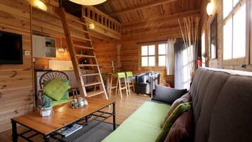 A wooden cabin interior with a sofa, a dining table, a ladder, and a television.