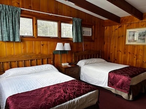 A wooden-paneled room with two beds, a nightstand with lamps, and a window with curtains.