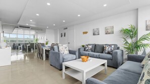 Ein modernes Wohnzimmer mit grauen Sofas, einem weißen Couchtisch und einem Essbereich, der durch eine Glaswand getrennt ist.