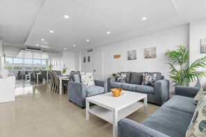 A modern living room with grey sofas, a white coffee table, and a dining area with a glass partition. - Oceanfront Family Retreat 2BR2BA Penthouse Pool & Parking (Carolina)