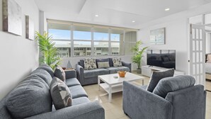 Ein modernes Wohnzimmer mit grauen Sofas, einem Couchtisch und einem Fernseher. Große Fenster und Topfpflanzen sind ebenfalls vorhanden.